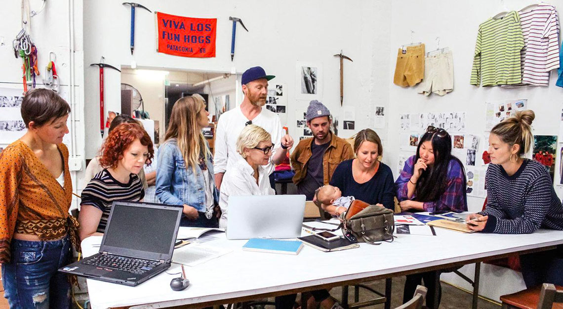 A group of 10 people in a workshop setting gathered around a table, a woman is holding a baby 