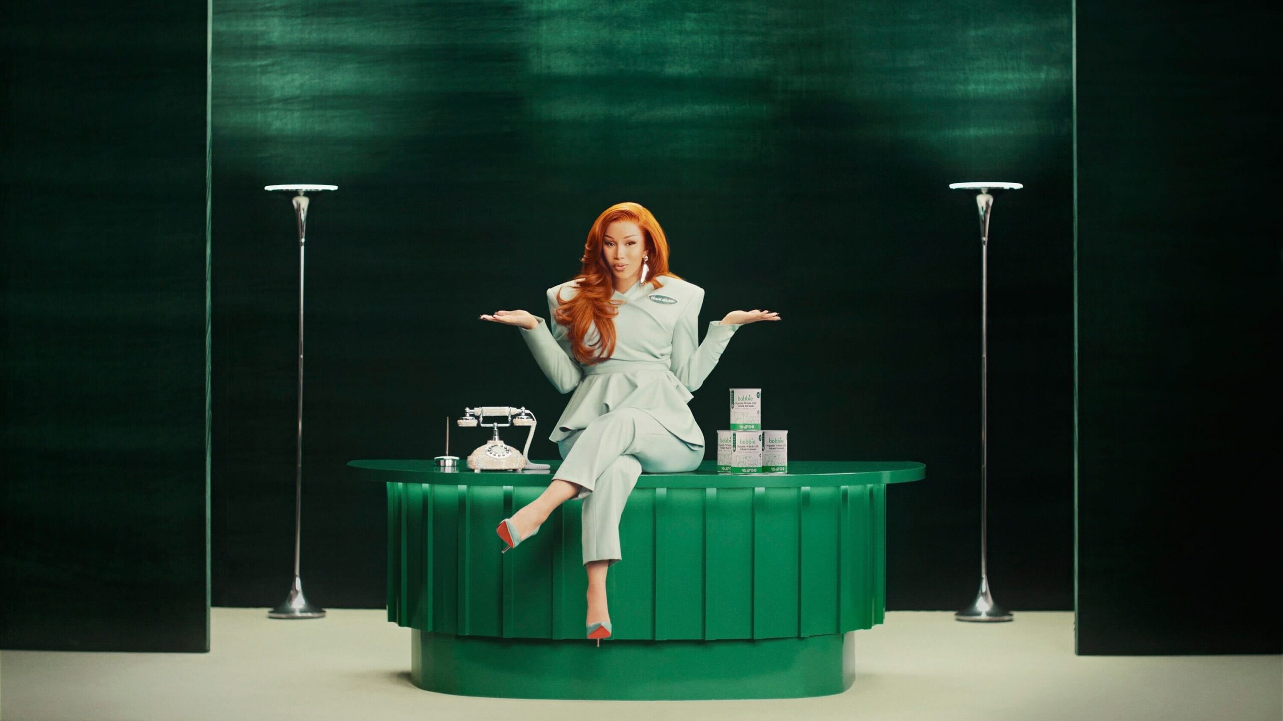 Cardi B sits on a desk, shrugging.